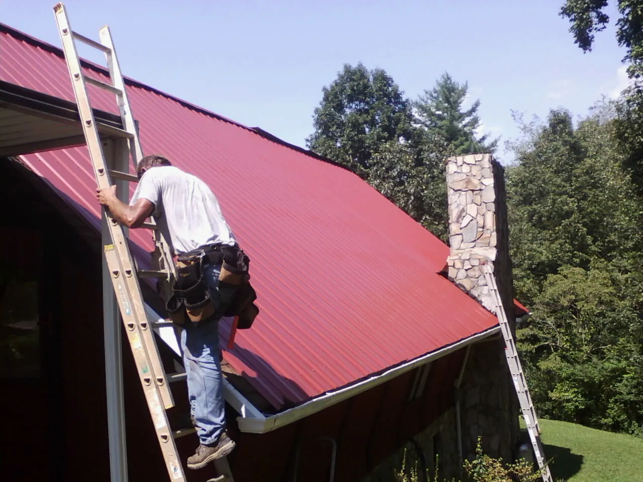 Roofer on a ladder beside a red metal roof and stone chimney — Soffit & Fascia Repair by Roofing Brothers in Braddyville